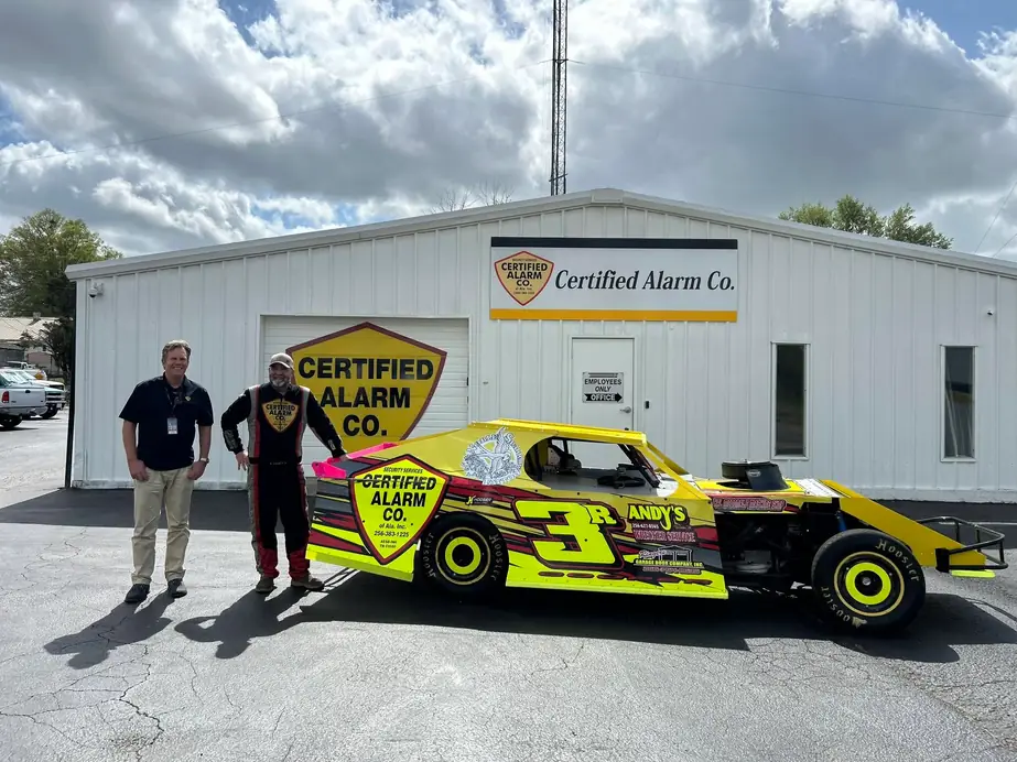 Two of the Certified Alarm crew beside the company's yellow number 3 dirt-track car, parked in front of the shop